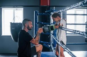 Conception of assistance. Young tattooed boxing coach teaches cute little girl in the gym photo