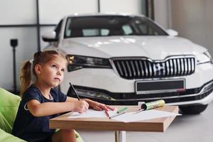 Cute little girl sits on the soft green chair by the table with pencil and paper sheets. Near modern automobile photo