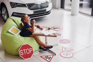 Cute little girl sits on the soft green chair by the table with pencil and paper sheets. Near modern automobile and road signs on the floor photo