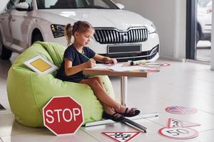 Cute little girl sits on the soft green chair by the table with pencil and paper sheets. Near modern automobile and road signs on the floor photo