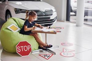 una linda niña se sienta en la suave silla verde junto a la mesa con lápiz y hojas de papel. cerca de automóviles modernos y señales de tráfico en el suelo foto