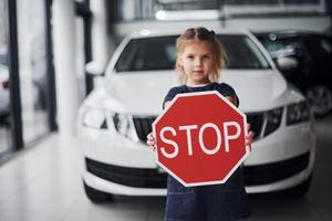 Portrait of cute little girl that holds road sign in hands in automobile salon photo
