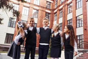 divirtiéndose y abrazándose unos a otros. grupo de niños con uniforme escolar que están juntos al aire libre cerca del edificio de educación foto