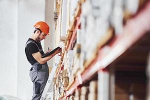 Storage worker stands on the ladder in uniform and notepad in hands and checks production photo