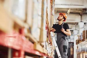 Storage worker stands on the ladder in uniform and notepad in hands and checks production photo