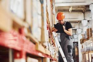 Storage worker stands on the ladder in uniform and notepad in hands and checks production photo