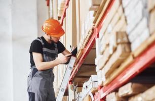 Storage worker stands on the ladder in uniform and notepad in hands and checks production photo