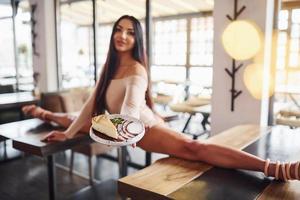 Young brunette in sexy wear soing stretches by sitting on the table and holds plate with food in restaurant photo