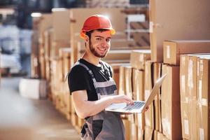 Storage worker in uniform and modern laptop in hands checks production photo