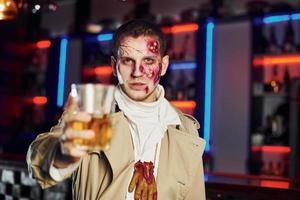 con bebida en la mano. retrato de hombre que está en la fiesta temática de halloween con maquillaje y disfraz de zombi foto