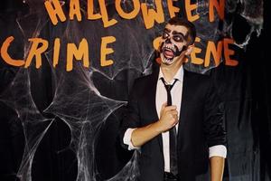 Portrait of man that is on the thematic halloween party in scary skeleton makeup and costume against crime scene wall photo