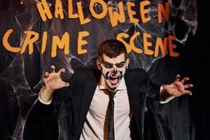 Portrait of man that is on the thematic halloween party in scary skeleton makeup and costume against crime scene wall photo