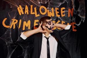 Portrait of man that is on the thematic halloween party in scary skeleton makeup and costume against crime scene wall photo