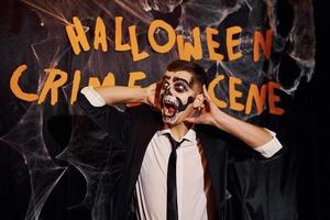 Portrait of man that is on the thematic halloween party in scary skeleton makeup and costume against crime scene wall photo