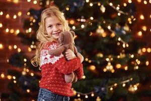 Cute little girl in red festive sweater with teddy bear indoors celebrating new year and christmas holidays photo