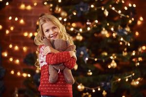 Cute little girl in red festive sweater with teddy bear indoors celebrating new year and christmas holidays photo