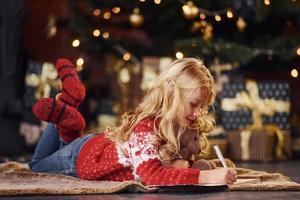 Cute little girl in red festive sweater lying down with teddy bear indoors celebrating new year photo