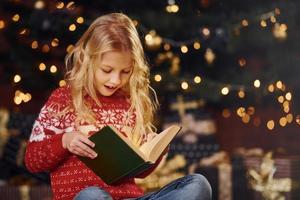 linda niñita con suéter festivo rojo leyendo un libro en el interior en las vacaciones de Navidad foto