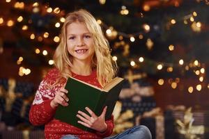 linda niñita con suéter festivo rojo leyendo un libro en el interior en las vacaciones de Navidad foto