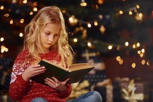 linda niñita con suéter festivo rojo leyendo un libro en el interior en las vacaciones de navidad foto