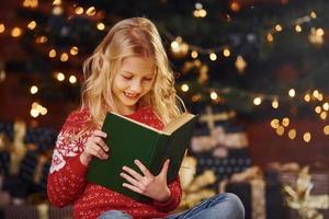 linda niñita con suéter festivo rojo leyendo un libro en el interior en las vacaciones de Navidad foto