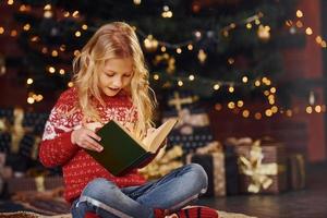 linda niñita con suéter festivo rojo leyendo un libro en el interior en las vacaciones de navidad foto