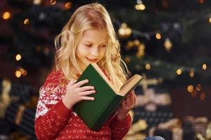 linda niñita con suéter festivo rojo leyendo un libro en el interior en las vacaciones de navidad foto