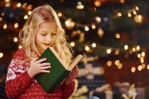 linda niñita con suéter festivo rojo leyendo un libro en el interior en las vacaciones de navidad foto