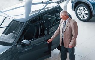 anciano con ropa formal caminando en el salón del automóvil y buscando el auto foto