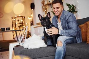 hombre sonriente en jeans sentado en el sofá en casa con su lindo perro foto