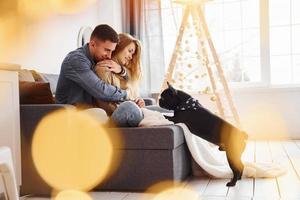 Happy young modern couple sitting on the sofa at home with christmas tree with their cute dog photo