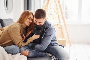 Happy young modern couple sitting on the sofa at home with christmas tree with their cute dog photo