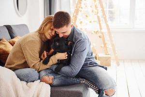 Happy young modern couple sitting on the sofa at home with christmas tree with their cute dog photo