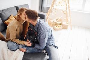 feliz joven pareja moderna sentada en el sofá en casa con árbol de navidad con su lindo perro foto