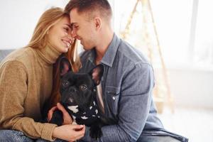 Happy young modern couple sitting on the sofa at home with christmas tree with their cute dog photo