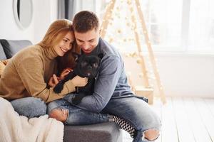 Happy young modern couple sitting on the sofa at home with christmas tree with their cute dog photo