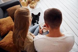 Happy young modern couple sitting on the sofa and writing wishes on notepad at home with their cute dog photo