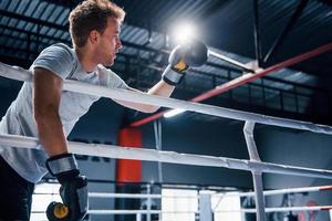 Tired young man in white shirt and protective gloves leaning on knots of boxing ring photo