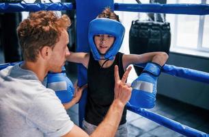 el joven entrenador de boxeo está ayudando a un niño con ropa protectora en el ring entre las rondas foto