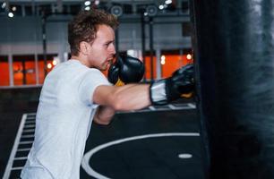 Young man in white shirt and boxing protective gloves doing exercises in gym with pushing bag photo