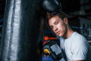 boxeador joven cansado en camisa blanca y con guantes protectores apoyado en saco de boxeo en el gimnasio foto