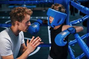 Young boxing coach is helping little boy in protective wear on the ring between the rounds photo