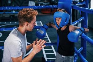 Young boxing coach is helping little boy in protective wear on the ring between the rounds photo