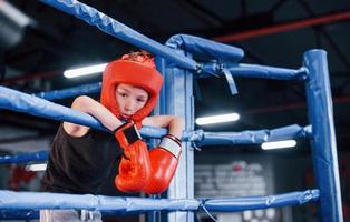 niño cansado en equipo de protección apoyado en los nudos del ring de boxeo foto