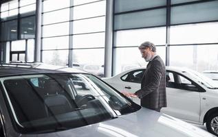 Senior businessman in suit and tie with gray hair and beard walking in auto salon and looking for new car photo