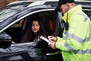 oficial de policía masculino en uniforme verde con bloc de notas revisando el vehículo en la carretera foto