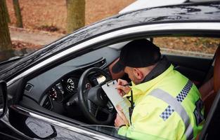 oficial de policía masculino con uniforme verde sentado en un automóvil y trabajando con documentos foto