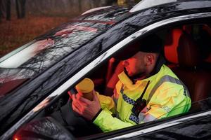 Male police officer in green uniform sitting in automobile with cup of drink photo