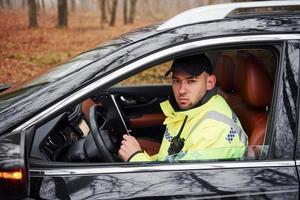 oficial de policía masculino con uniforme verde sentado en un automóvil y trabajando con documentos foto