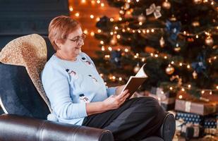 Grandmother sitting on the chair with book in festive New year room with christmas decorations photo